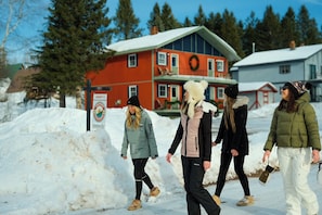Un groupe de personnes se promène dans un endroit enneigé, près d’un grand tas de neige, d’un bâtiment rouge orné d’une guirlande et d’un panneau avec un logo circulaire.