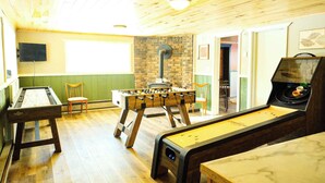 A room with a foosball table, a pool table, and a shuffleboard table. There is a brick wall and a television mounted on the wall. - Elevated Ski Chalet at Powderhorn Mountain (Bessemer)