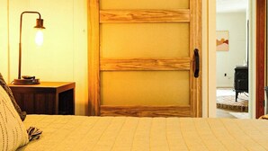 Un dormitorio con una cama, una mesita de noche con lámpara, una puerta corredera de madera y vistas a otra habitación.