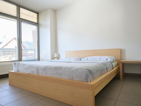 A minimalist bedroom with a wooden bed, a bedside table, and a window with blinds.