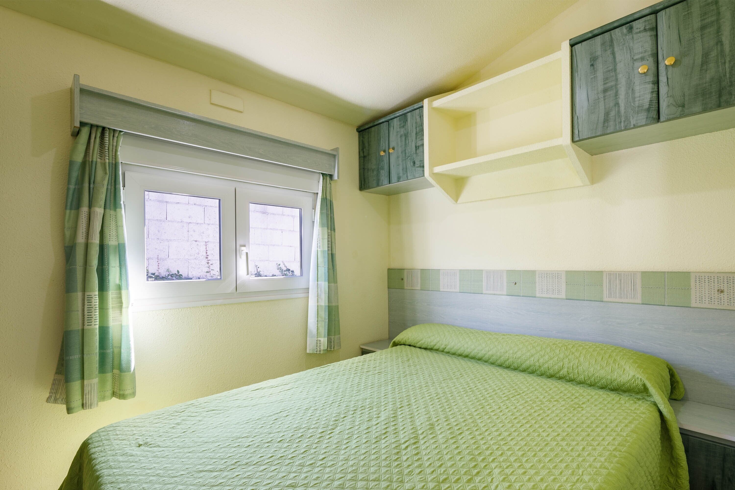 A room with a bed, a window with curtains, a wall-mounted shelf, and a headboard.