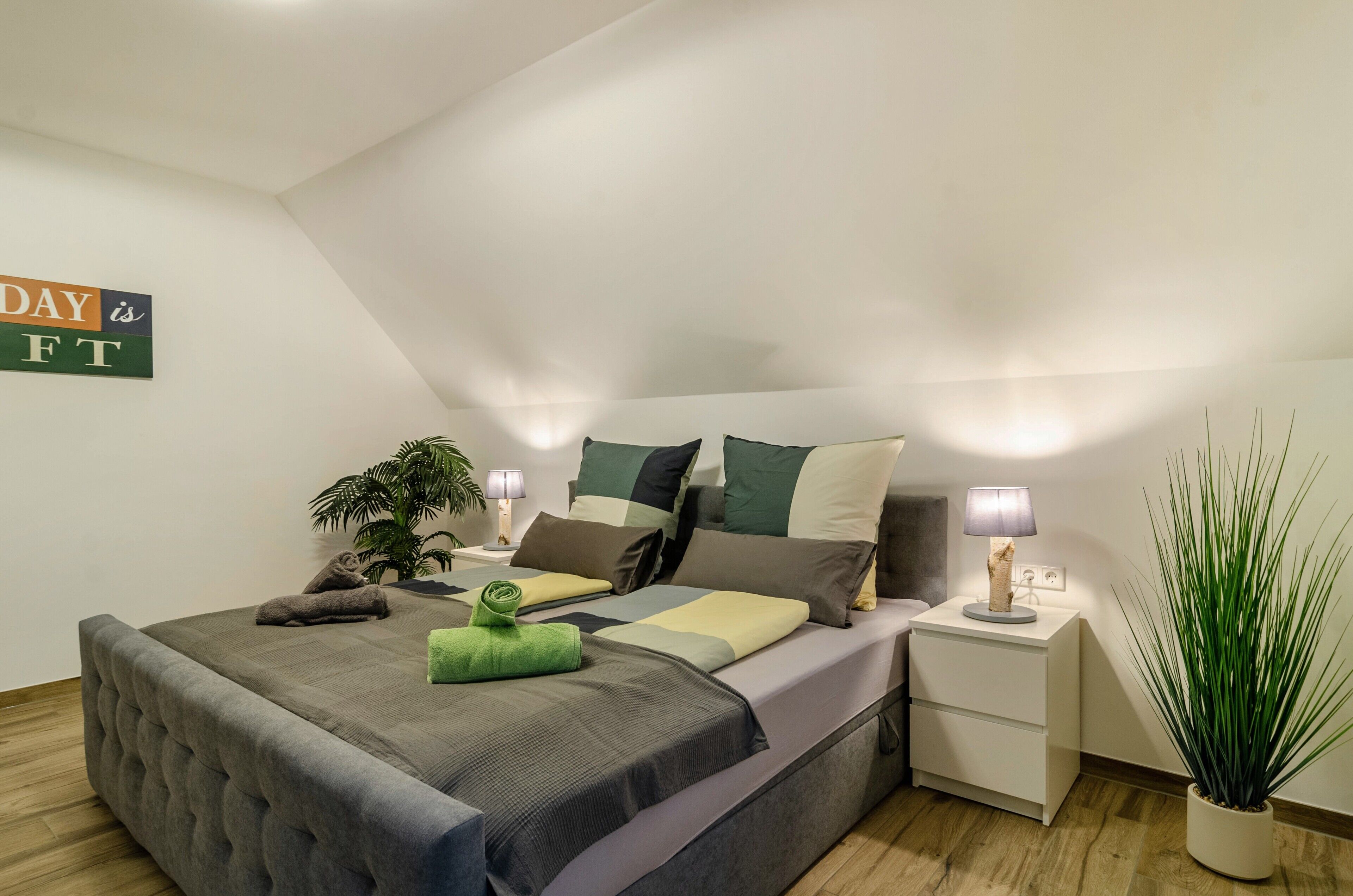 A bedroom with a bed, nightstands, a lamp, a potted plant, and a wall sign.
