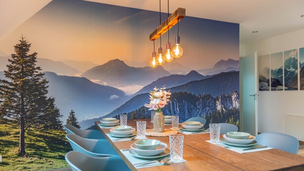 A dining area with a wooden table set for four, blue chairs, a mountain view mural, and a hanging light fixture. - Apartment 'Bergblick' with Shared Terrace, Sauna, Whirlpool & Pool (Stockach)