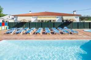 A pool area with sun loungers and a building with a red-tiled roof and awnings.