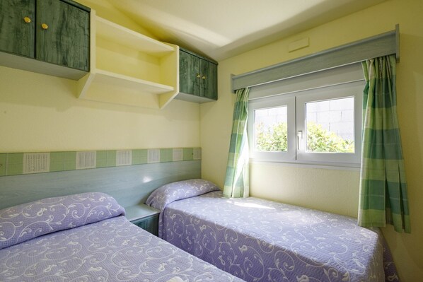 A room with two beds, a window with curtains, a shelf unit, and a headboard with a patterned design.