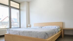 Quarto com cama de madeira, mesa de cabeceira, janela com persianas e radiador.