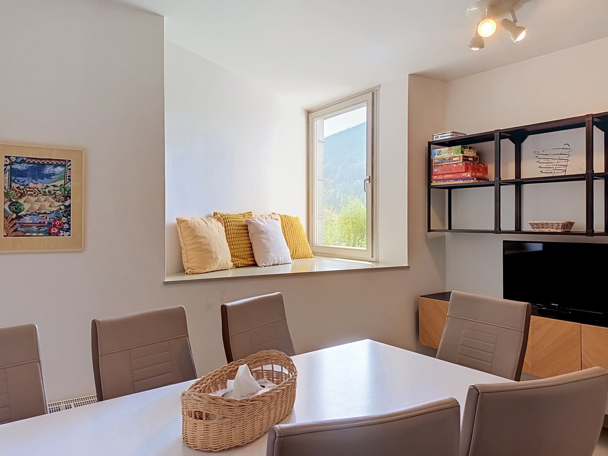 A dining area with a table, chairs, a window with pillows, a bookshelf, and a framed picture on the wall.