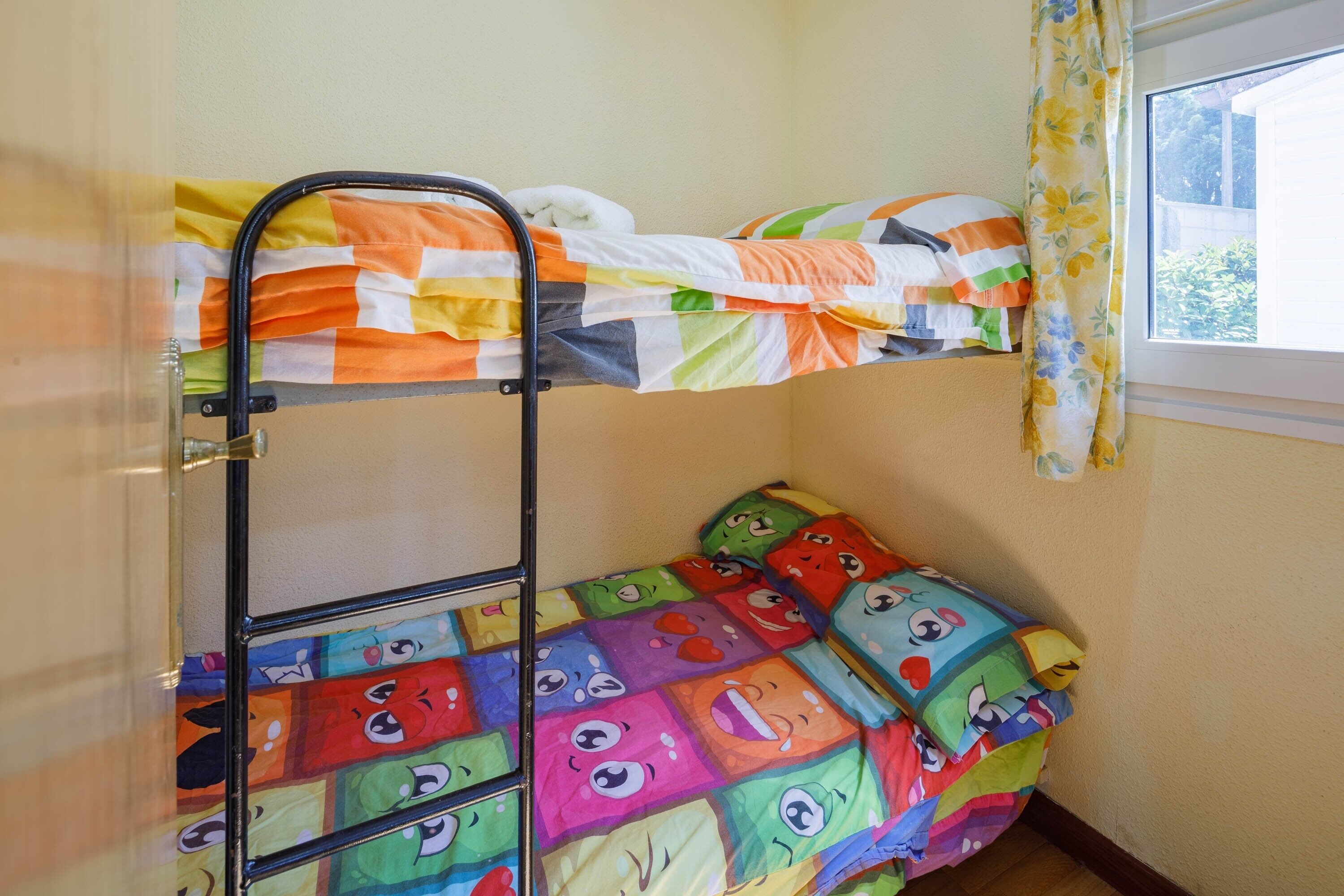 Una cama litera con ropa de cama colorida, una ventana con cortinas y una pared sencilla.