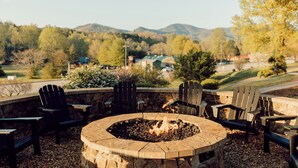 An outdoor seating area with a stone fire pit and Adirondack chairs, surrounded by a stone wall and a view of a residential area and hills.