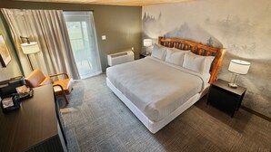 A hotel room with a large bed, a desk with a chair, a nightstand with a lamp, and a wall with a forest mural.