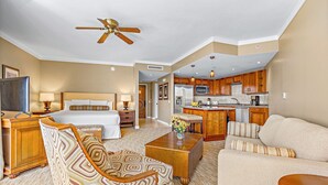 A hotel room with a bed, a desk, a chair, a sofa, a coffee table, a TV, a ceiling fan, and a kitchen area with a microwave, refrigerator, and sink.