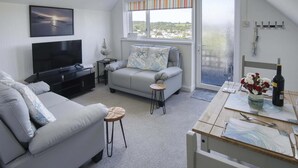 Un salón moderno con un sofá gris, un televisor, una mesita con una botella y flores, y una ventana con vistas a la ciudad.