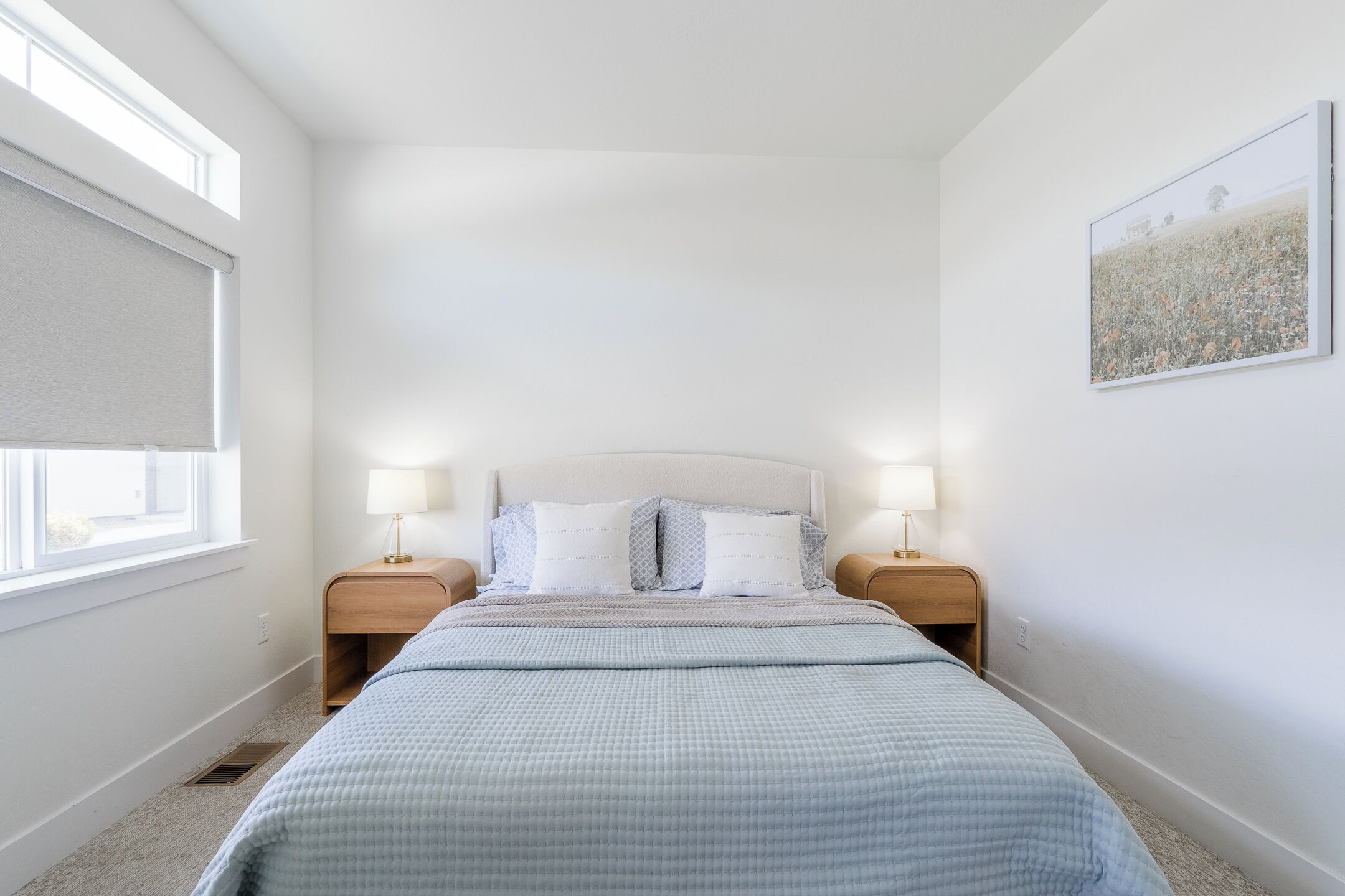 A bedroom with a large bed, two bedside tables with lamps, a window with a blind, and a framed picture on the wall.