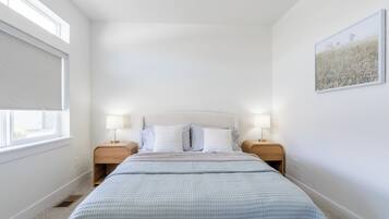 A bedroom with a large bed, two bedside tables with lamps, a window with a blind, and a framed picture on the wall.