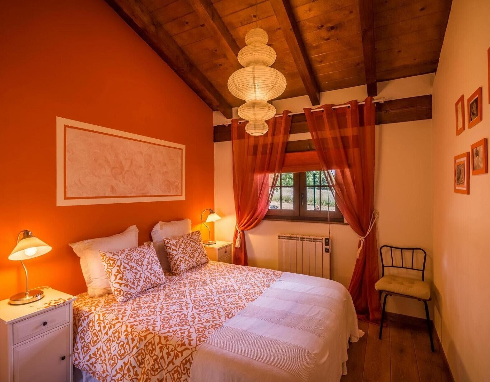 A bedroom with a bed, two bedside lamps, a radiator, a chair, and a window with curtains.
