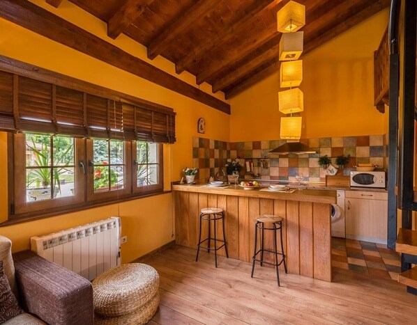 Una cocina con vigas de madera, salpicadero a cuadros, barra con taburetes y ventana con persianas.