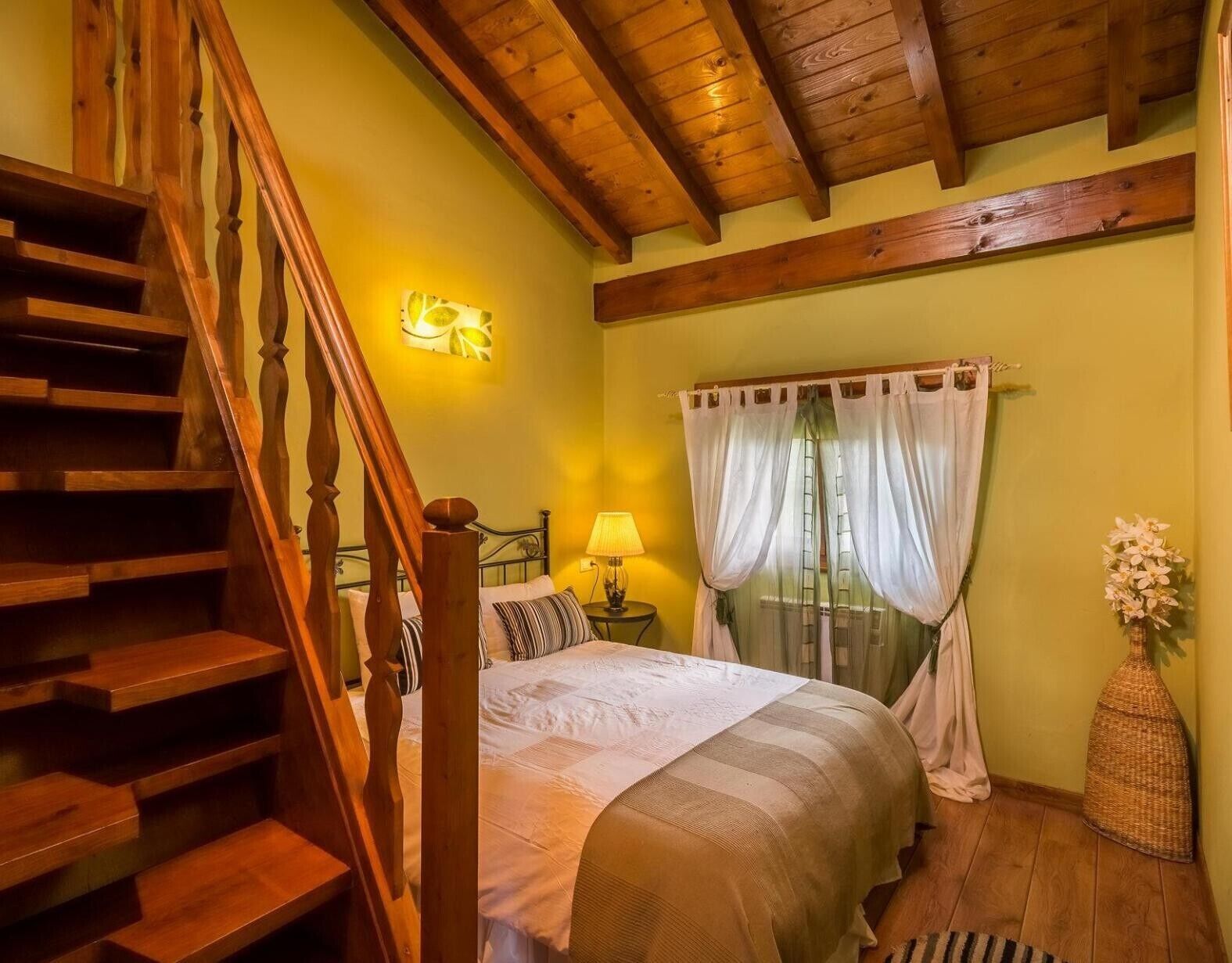 Un dormitorio con una escalera de madera, una cama con colcha blanca y a rayas, una mesita de noche con lámpara y una ventana con cortinas translúcidas.