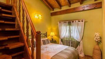 Un dormitorio con una escalera de madera, una cama con colcha blanca y a rayas, una mesita de noche con lámpara y una ventana con cortinas translúcidas.