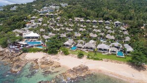 An aerial view of a resort with multiple bungalow-style accommodations, a central pool area, and a beach.