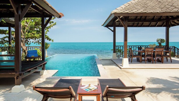 A poolside area with a view of the sea, wooden furniture, and a covered seating area.