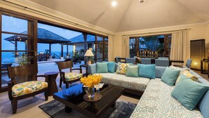 A spacious living area with a large sofa, chairs, and a coffee table. There is a view of the ocean through large windows.