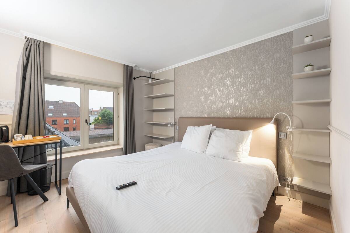 Hotelkamer met een groot bed, een bureau met stoel, een raam met gordijnen en inbouwplanken.