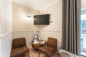 Egy sarokszobában két barna bőr fotel, egy kis, fából készült asztal, rajta egy ventilátor, és a falra szerelt televízió található.