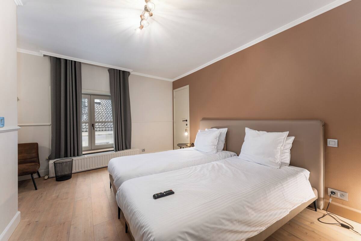 Een hotelkamer met twee bedden, een raam met gordijnen, een radiator, een stoel en een nachtkastje met een lamp.