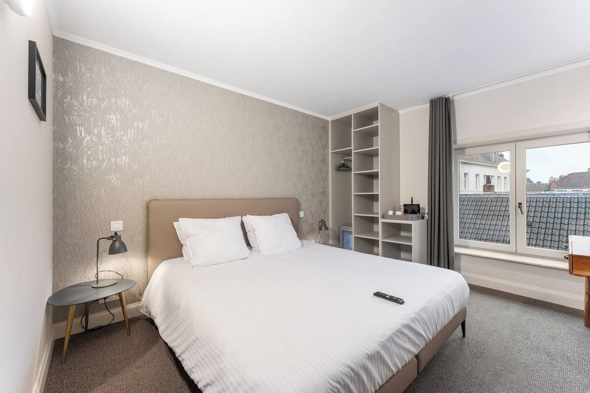Hotelkamer met een groot bed, nachtkastje met lamp, kast en uitzicht op de stad door een raam.