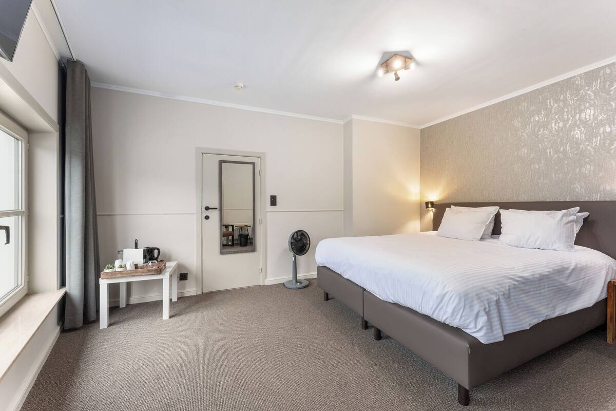 Een hotelkamer met een groot bed, een nachtkastje, een klein tafeltje met een dienblad, een ventilator en een raam met gordijnen.
