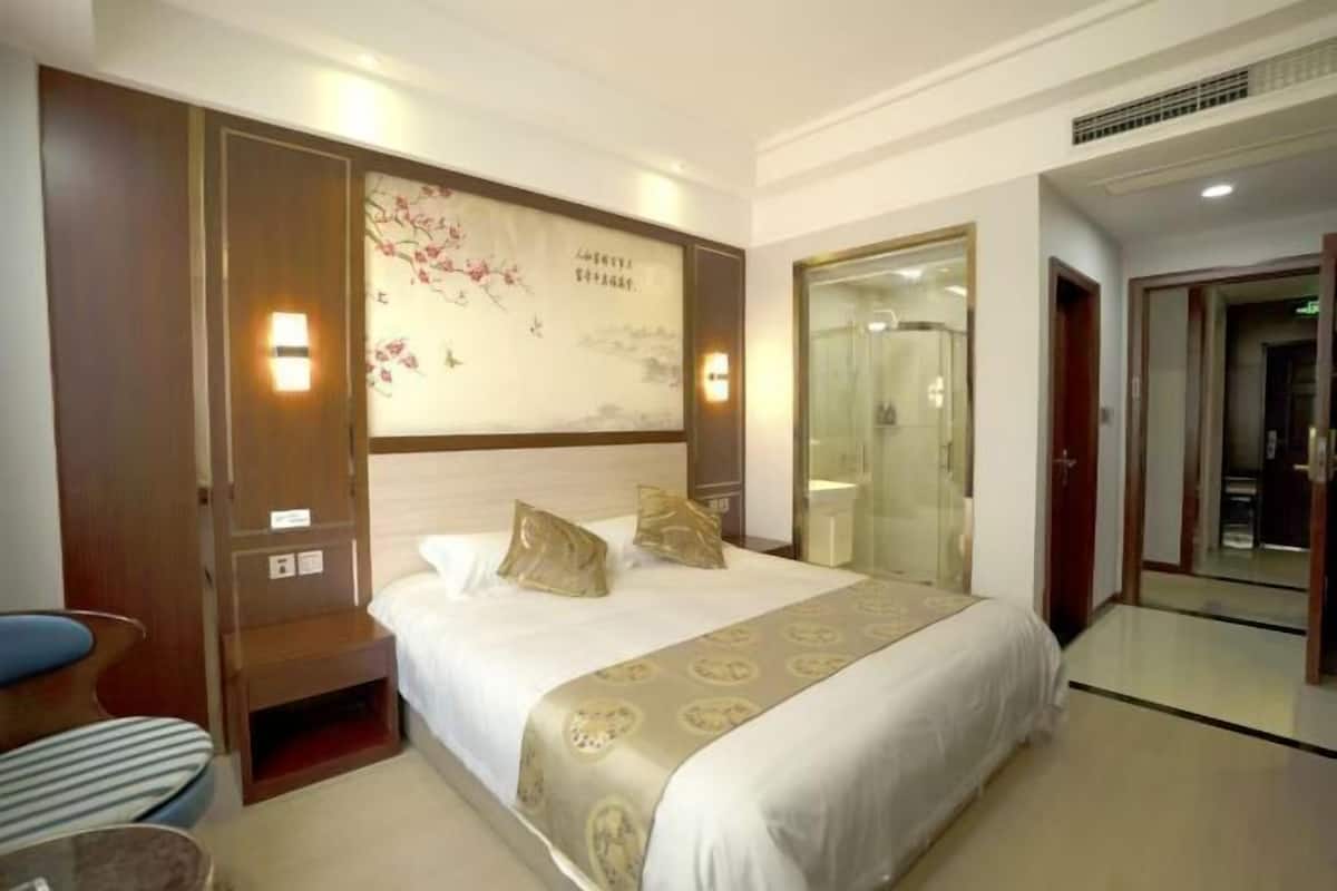 A hotel room with a bed, a bedside table, a chair, a wall with a decorative painting, and a bathroom door.