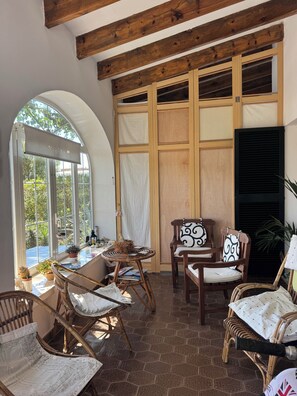 A room with a large arched window, wooden ceiling beams, wicker furniture, and a tiled floor.