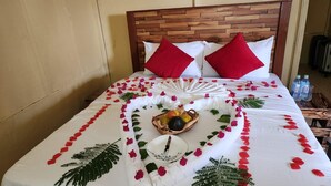 Una cama bien hecha con cabecera de madera, ropa de cama blanca, almohadas rojas y un arreglo decorativo de pétalos de rosa y una cesta de frutas.