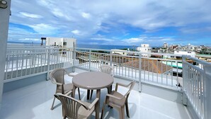 A balcony with a round table and four chairs, overlooking a cityscape and the sea. - Palace Oceanside (Chatan)