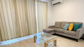 A modern living room with a glass coffee table, a beige sofa with colorful cushions, and a large window with sheer curtains. - Palace Oceanside (Chatan)