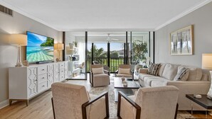 A spacious living room with a large flat-screen TV, comfortable seating, and a view of a tropical landscape through large windows. - Beach Life 7622 (Fort Pierce)