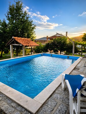 Une piscine avec une terrasse carrelée, un espace couvert et une vue sur une maison au loin.