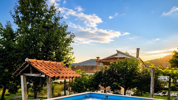 Ein Schwimmbecken mit gefliestem Deck, einem überdachten Sitzbereich und Blick auf ein Haus und Hügel im Hintergrund.