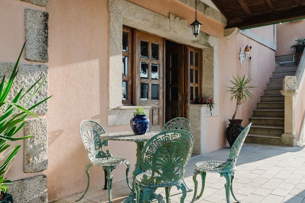 Un bâtiment en pierre avec une porte en bois, une fenêtre et un escalier. On y trouve deux chaises en métal vertes et une table sur laquelle est posé un vase bleu.