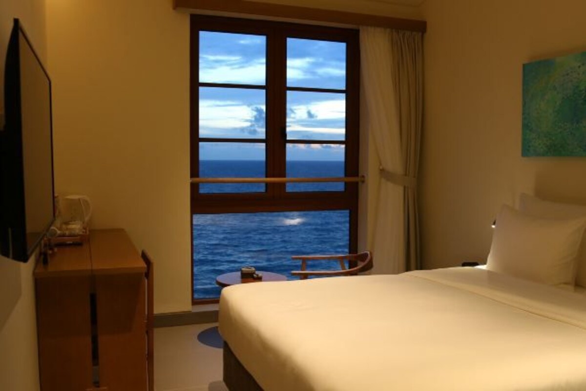 A hotel room with a bed, a small table, a chair, a TV, and a window with a view of the sea.