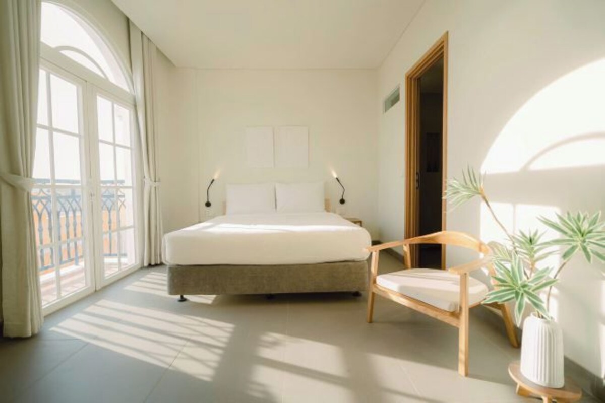 A bedroom with a bed, a chair, a bedside table, and a potted plant.