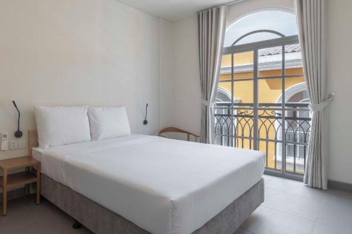 A bedroom with a bed, a bedside table, a lamp, and a window with a view of a yellow building and a balcony.