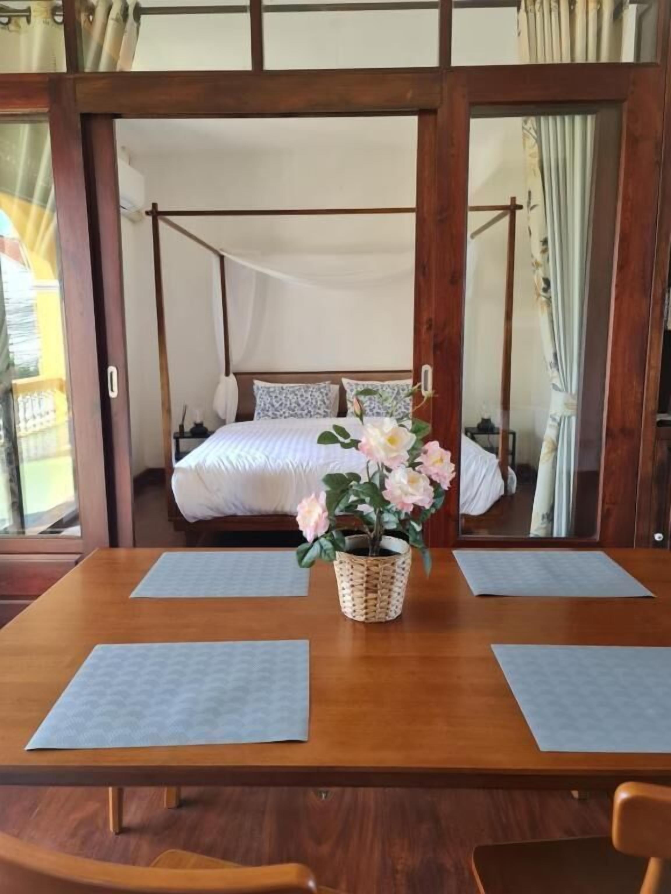 Una habitación tradicional japonesa con puertas correderas de madera, una cama con dosel y una pequeña mesa con una cesta tejida y un jarrón con flores.