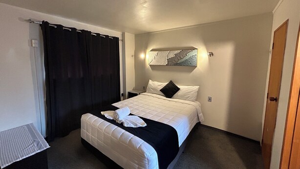 A hotel room with a bed, a nightstand, a black curtain, a wall-mounted artwork, and a door. - Aquarious motor inn (Hamilton)