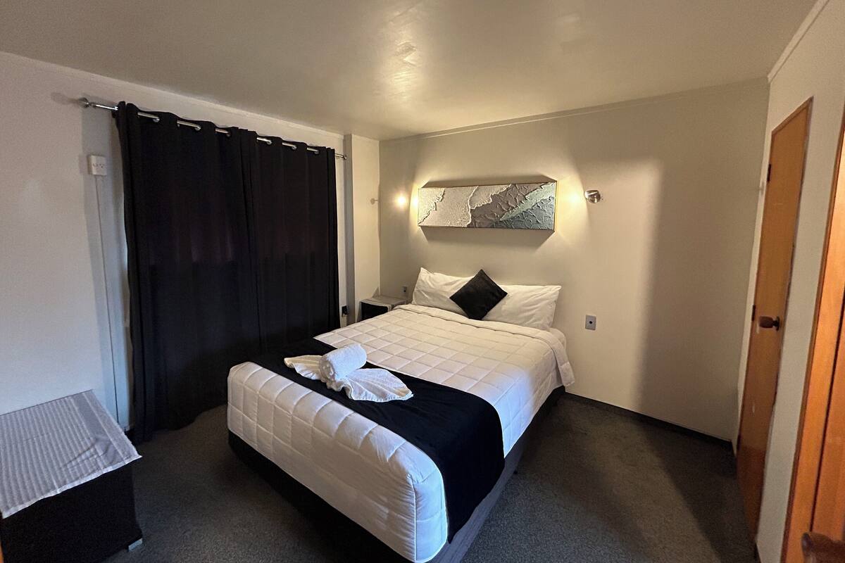 Une chambre d’hôtel avec un lit, une table de chevet, un rideau noir, une œuvre d’art accrochée au mur et une porte.