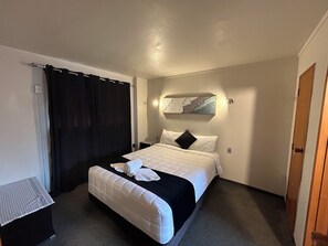 A hotel room with a bed, a nightstand, a black curtain, a wall-mounted artwork, and a door. - Aquarious motor inn (Hamilton)