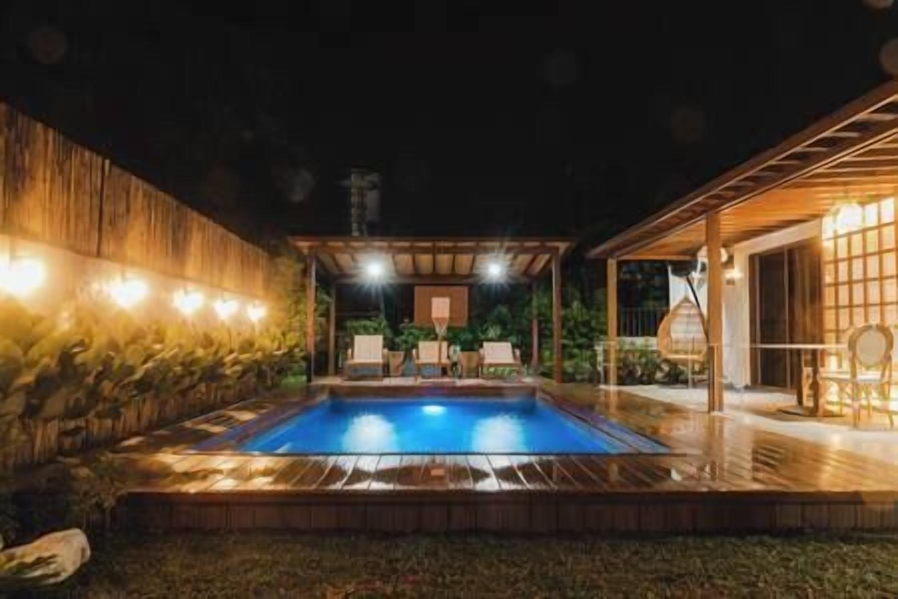 Piscina com deck de madeira, área de estar coberta e estrutura de vidro com cadeiras.