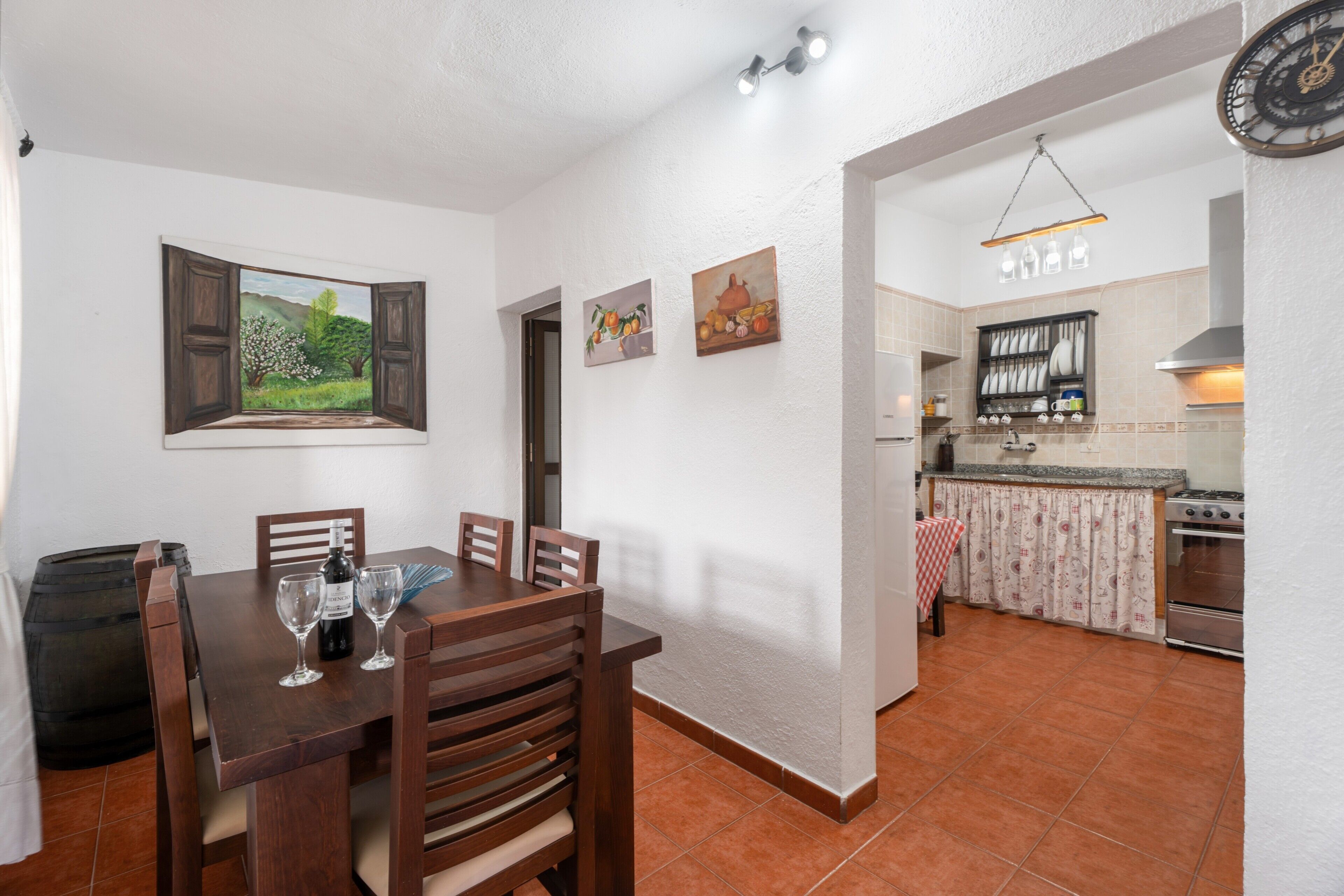 Un coin repas avec une table en bois, des chaises et une bouteille de vin. On aperçoit une cuisine à l’arrière-plan, avec une cuisinière et des placards.