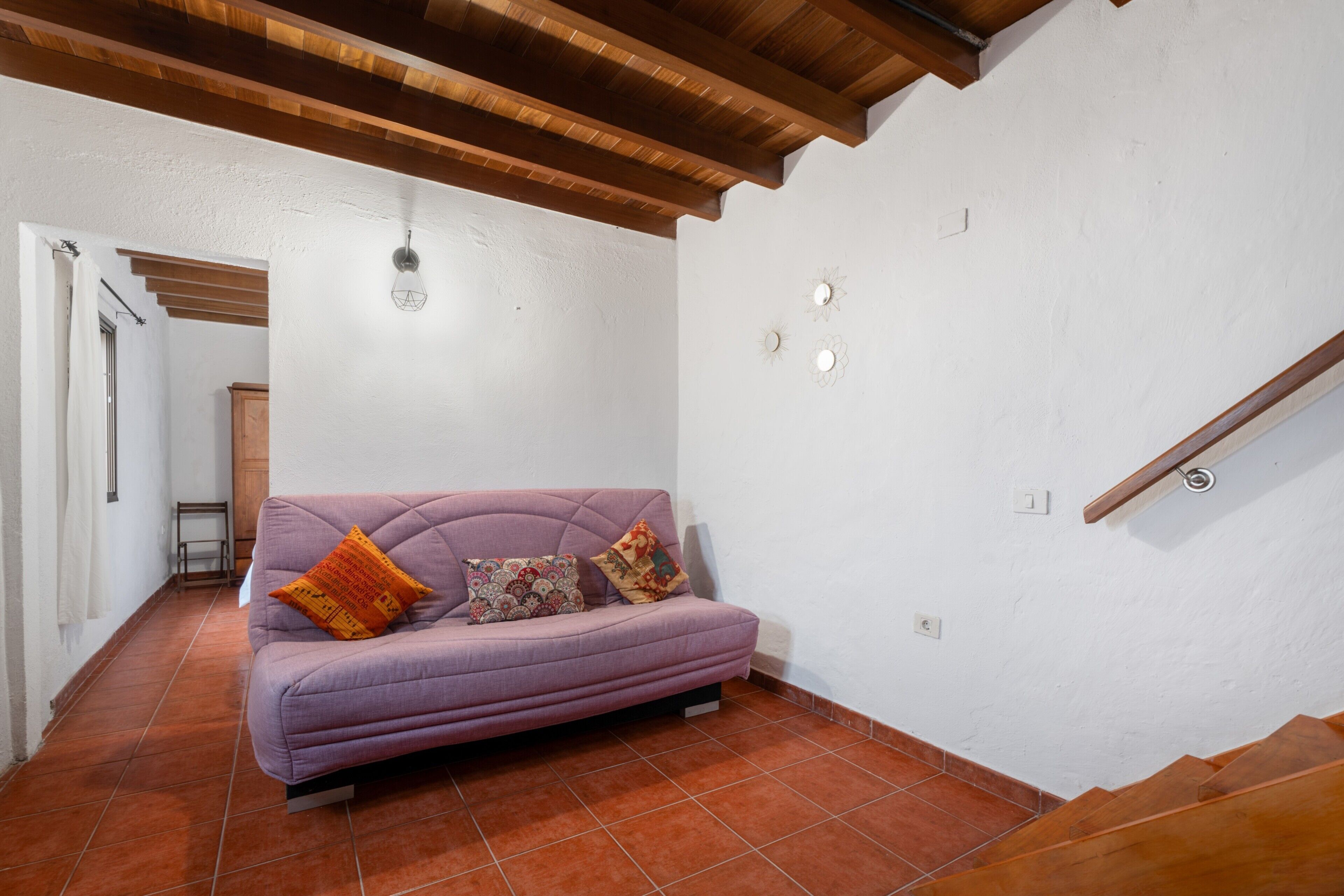 A room with a purple sofa, wooden ceiling beams, white walls, and a tiled floor.