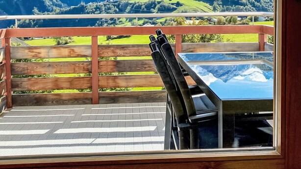 Un terrazzo in legno con tavolo e sedie, affacciato su un paesaggio montuoso sotto un cielo sereno.